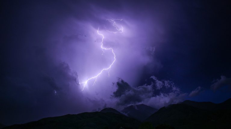夜間雷暴中劃破紫色烏雲的巨大閃電，下方為黑暗的山脈輪廓。